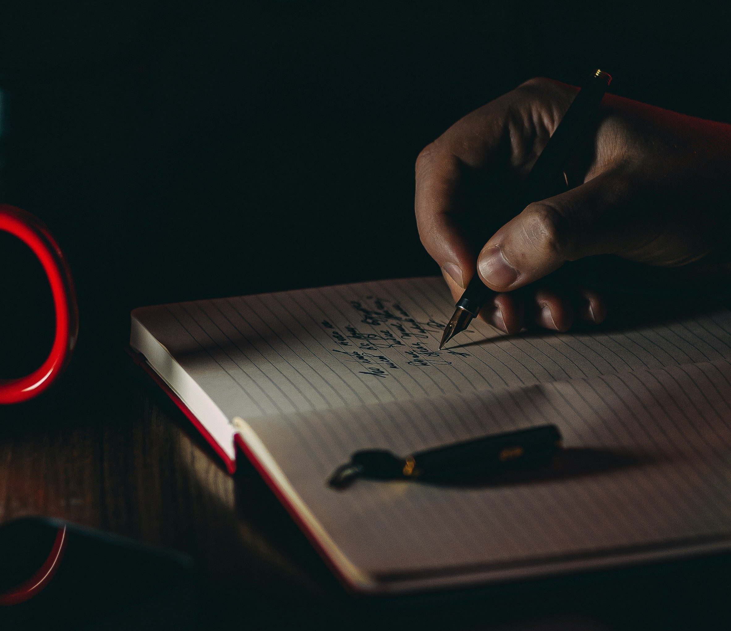 A hand writing with a fountain pen in a notebook, lit by a small warm lamp